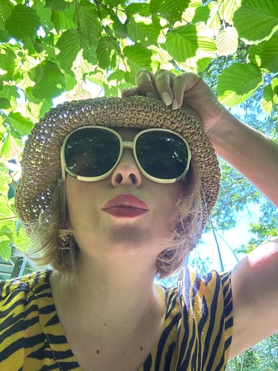 In Sochi, Russia, under the dappled sunlight filtering through green leaves, a woman is captured in a close-up, low-angle, outdoor selfie. She is wearing a straw hat and large, light-rimmed sunglasses, with her right hand raised to adjust the hat. Her lips are painted a soft red, and she appears to be looking directly into the camera. She has blonde hair and is wearing a yellow and black striped garment. The background is a blur of green foliage and bright blue sky, suggesting a natural, possibly park-like setting.