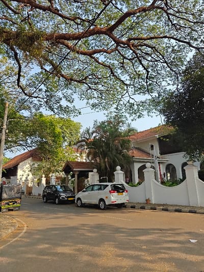 The image displays a street scene in Kochi, India, under a clear sky partially covered by a dense, leafy tree canopy. The foreground features an asphalt road with visible shadows. A white Toyota Innova multi-purpose vehicle (MPV), marked "TAXI" with license plate KL 07 BZ 3330, is parked parallel to the right curb, facing away from the viewer. A black Toyota Innova Crysta SUV is positioned slightly ahead of the white MPV, also facing away. On the far left, a dark-colored three-wheeled auto-rickshaw is partially visible.
A white, decorative boundary wall with pillars topped by spherical finials and curved sections separates the road from a large, white building with a red-tiled roof. This building features multiple arched openings and a prominent dark-wood, tiled portico above an entrance. A white flag, displaying a blue emblem, is affixed to the building's façade. Lush green vegetation, including palm trees, is visible within the property behind the wall. A utility pole stands on the far left, supporting overhead wires. A small terracotta pot rests near the base of the white wall on the right. No individuals are visible.