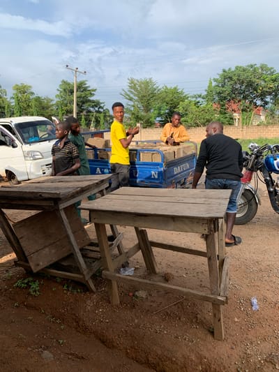 The image shows a street scene in Kabba, Nigeria. A blue tricycle with the word "DAYUN" on the side is partially visible, loaded with rectangular objects that appear to be concrete blocks. A man in a long-sleeved orange shirt sits on top of the load. Another man, wearing a black long-sleeved shirt and blue jeans shorts, stands at the back of the tricycle, seemingly observing. Three young boys are near a white van, one in a yellow shirt holding up his hands, another wearing a green shirt leaning over the side, and a third wearing a striped shirt. In the foreground stands a rough, wooden table or counter made of unfinished planks. A motorcycle is partially visible at the top of the image. The ground is dirt. Trees and buildings are visible in the background.