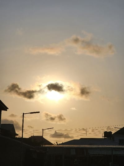This image captures an urban landscape in Lekki, Nigeria, at either sunrise or sunset. The sun is centrally positioned in the upper mid-ground, partially obscured by dark, patchy clouds, radiating a strong light that transitions the sky from bright white-yellow around the sun to a pale blue-grey towards the top. Several smaller, diffused clouds are scattered across the sky.

The lower third of the image features a dark, jagged silhouette of multiple building rooftops, indicating a developed area. Two distinct streetlights or utility poles with horizontal fixtures are prominently silhouetted against the bright sky, one larger on the left and a smaller one further right. Numerous parallel utility wires extend horizontally across the mid-ground, connecting unseen poles. On one of the distant rooftops, two dark, cylindrical objects, likely water tanks, are visible. The immediate foreground is dominated by a dark, solid fence or wall, obscuring any ground-level details. No individuals or specific actions are discernible within the frame.