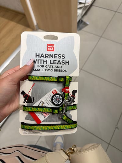 A person's hand with a light pink manicure is holding a packaged cat and small dog harness with a leash. The product packaging is white and features a cartoon illustration of a cat and a dog. The harness and leash are green with a yellow and black floral pattern and are attached to the packaging. A red tag with white text that reads "WAU CAT" is visible. A QR code is also present on the packaging, along with instructions for scanning and registering the product. The background appears to be a retail environment with tiled flooring and a light-colored wall, suggesting the image was taken in a store. The presence of a light-colored wall and tiled floor is consistent with many retail spaces found in Sofia, Bulgaria.