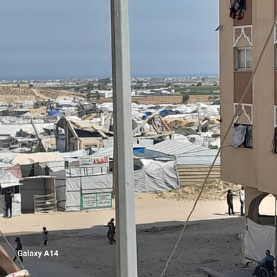 The image, taken during the daytime under a clear sky, depicts a densely populated area in what appears to be a displacement or refugee camp, consistent with the Khan Yunis context.
In the foreground, a multi-story building with light brown and darker brown sections and decorative white window frames stands on the left. Clothes are seen hanging to dry from one of its windows. Several thin wires or cables crisscross the frame.
The mid-ground and background are dominated by a sprawling settlement of numerous makeshift shelters. These structures are constructed from various materials, including white, grey, and beige tarpaulins, wood, and possibly corrugated metal, indicating temporary and improvised living conditions. They are packed closely together, extending far into the distance towards the horizon.
A dirt or unpaved path runs along the right side of the image, where several individuals are visible. One person, wearing a blue top and dark trousers, is pushing what appears to be a wheelchair or cart. Further down the path, two darker figures are walking. Other figures can be seen near the entrance of the permanent building.
Arabic text is visible on some of the banners and signs within the camp. One sign with a green border and text on a white background, potentially reading "ليون حلويات" (Lyon Sweets), and another prominent sign with red Arabic text, possibly "المجد" (Al-Majd, meaning "The Glory"), can be identified. The watermark "Galaxy A14" is present in the bottom right corner of the image.