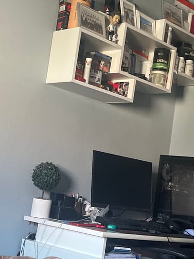 This scene is located in Pokrovnik, Bulgaria.
The image captures an indoor environment, likely a personal room or workspace, featuring a light grey textured wall. A white, multi-tiered floating shelf system is mounted on the upper section of the wall. The shelves hold a variety of personal items: multiple framed photographs, including group portraits and individual or couple shots; a bobblehead figurine resembling Lionel Messi in an Argentina soccer jersey; and several containers of dietary supplements, specifically a large black tub labeled "CREATINE" with green text, and other containers partially visible with labels such as "WHEY" and "BEST WHEY." A rolled United States $100 bill is placed beneath the creatine container. Additional boxes, including one marked "Unic" and another "Everyday balan...", and various bottles are also on the shelves.
Below the shelving unit, a white desk or table surface is equipped with computer hardware and accessories. On the left, a small, artificial topiary plant in a white cylindrical pot is present. Adjacent to it are black desk organizers holding pens and pencils. Two dark computer monitors are positioned on the desk, accompanied by a black keyboard, a white computer mouse, and a pair of black headphones. A small green object rests on the keyboard. To the right, a desktop computer tower with a transparent side panel displays its internal components. Numerous cables and wires are visible on and around the desk, indicative of connected electronics. A flat, red object is situated near the keyboard. The area beneath the desk is partially obscured but shows additional cables and possibly papers.