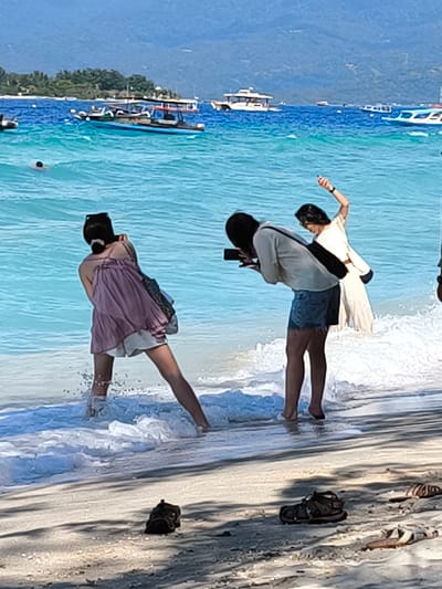 In a beach scene in Unknown, Indonesia, three individuals are present near the shoreline. One person, positioned in the foreground and facing away from the camera, stands in shallow water, their lower legs obscured by splashing waves. They wear a light-colored skirt and a darker top. To their right, another individual is actively taking a photograph with a smartphone. This person is wearing a light-colored shirt and denim shorts. Behind them, a third person in a cream-colored dress gestures with their right arm raised. The ocean is a vibrant turquoise blue, with several boats visible on the horizon. The beach in the foreground features scattered dark-colored sandals.