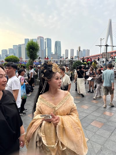 Here's a description of the image:
The image shows a woman in a traditional golden dress with ornate hair decorations holding a small electronic device, possibly a phone or camera, in a public setting. She's positioned in the foreground, slightly angled towards the camera. The background includes a crowd of people, and in the distance, a bridge with prominent cables and modern city buildings, indicating an urban environment. Based on the city skyline and bridge architecture, this image was likely taken in Yuzhong District, Chongqing, China, near the Dongshuimen Bridge. The weather appears overcast, as the sky is cloudy.