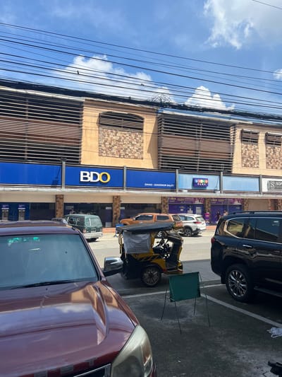The image shows a street view in Davao City, Philippines, featuring a BDO bank branch. The building has a modern facade with horizontal louvers and stone accents. A prominent blue banner displays the BDO logo. To the right of BDO, a "Bingo Plus" establishment is visible. Several vehicles are parked and on the street, including a maroon SUV in the foreground, a black SUV, a green van, and a yellow tricycle. People are seen walking in front of the businesses and in the background. The sky above is blue with scattered clouds, and electrical wires crisscross the scene. The overall environment suggests a commercial street on a sunny day.