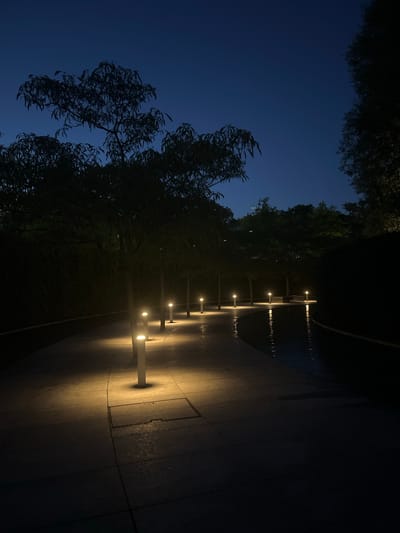 The image depicts a nighttime scene of an outdoor walkway lined with modern, low-profile bollard lights. The walkway appears to be paved with large, dark tiles, and it curves gently to the right, leading into darkness. The lights cast warm pools of illumination on the path, highlighting the textured surface. To the left of the walkway, dark silhouettes of trees with feathery foliage are visible against a deep blue twilight sky. A faint hint of a star is present in the upper left quadrant of the sky. The overall mood is serene and tranquil, suggesting a quiet evening in a park or public garden. The scene is illuminated by the artificial lights and the fading twilight, creating a dramatic contrast between light and shadow. There is no visible text in the image. The location context of Krasnodar, Russia, suggests this could be a public space within the city, designed for evening ambiance.