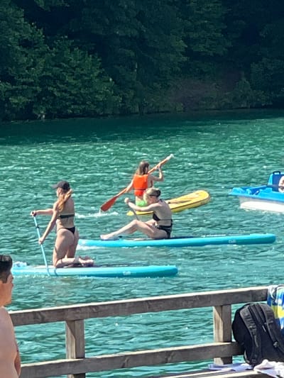 The image depicts a sunny day on a lake, with several people enjoying various water activities. In the foreground, a wooden railing separates the viewer from the water. Behind the railing, three individuals are visible on paddleboards. One person, wearing a black bikini and a black baseball cap, is kneeling on a blue paddleboard and holding a paddle. Another person, wearing a black bikini, is sitting on a blue paddleboard. Further in the background, a third person, wearing an orange life vest and green shorts, is paddling a yellow inflatable. A blue boat with a white hull is also present on the water. The lake is surrounded by lush green trees, suggesting a natural or park-like setting. The water is a vibrant turquoise color, reflecting the bright sunlight. The overall scene conveys a sense of leisure and outdoor recreation. This scene is likely at the Vingo Park's recreational lake in Vilnius, Lithuania, known for its water sports and leisure activities.