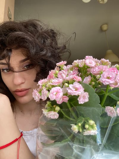 The image shows a close-up of a young woman holding a bouquet of pink flowers. She has dark, curly hair and is looking towards the camera with a neutral expression. A red bracelet is visible on her left arm. In the background, a ceiling light fixture and part of a wall are visible. The setting appears to be indoors, possibly a residential apartment, with a window or opening to the outside, indicated by the light and the presence of a ceiling fixture. There is no specific indication of the location being in Sofia, Bulgaria.