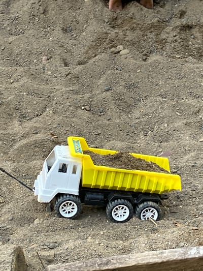 An overhead shot shows a toy dump truck filled with dirt. The truck is white and yellow and has six white wheels. The truck is positioned in a sandy area that appears to be a playground or construction site. There are some small rocks and debris scattered throughout the sand. The image does not contain any elements that can be used to infer a specific location in Lhokseumawe, Indonesia.