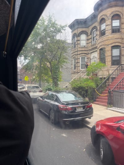 The image shows a street scene in what appears to be a residential area of New York, USA. In the foreground, the interior of a vehicle is visible, with a person's arm and shoulder covered in black fabric. The perspective suggests the photo was taken from inside this vehicle, looking out through the sunroof.
Outside, a row of brick apartment buildings with arched windows lines the street. A red car is parked to the right, and a black car is parked in front of it. A person wearing a dark tank top and shorts is leaning over a white railing on the stairs of one of the buildings. Yellow caution tape is strung across the stairs. Trees and greenery are present along the sidewalk and in front of the buildings.
Further down the street, a yellow school crossing sign is visible, indicating the presence of a school or a school zone. The sky appears overcast, suggesting a cloudy day. The overall impression is a snapshot of everyday life on a New York City street, possibly capturing a minor incident or construction work due to the caution tape.