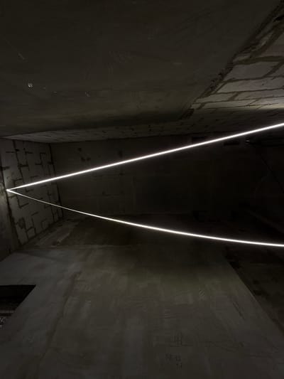 The image shows a dark, unfinished interior space with concrete walls and ceiling, reminiscent of a construction site or an underground area. Two bright, linear light fixtures are prominently featured, casting a stark glow across the scene. The walls are constructed from concrete blocks, some of which appear rough and unplastered. The floor is also concrete and shows signs of wear and unevenness. In the lower right quadrant of the image, some handwritten markings in what appears to be Cyrillic script are visible on the wall, reading "пв 1.6" and "1.7". The overall atmosphere is stark and industrial. This location is not identifiable as being in Moscow, Russia, based solely on the visual information provided.