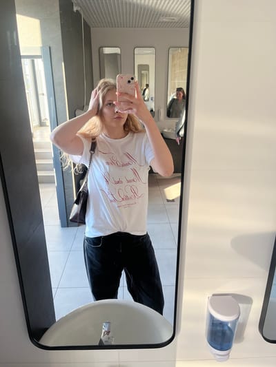 A young woman with blonde hair is taking a mirror selfie in a public restroom. She is wearing a white t-shirt with red script writing and black pants. She has her hands on her head and is looking at the camera. In the reflection of the mirror, another woman is visible in the background. The restroom has tiled walls and floors, several mirrors, and sinks. A soap dispenser is mounted on the wall. The scene appears to be set in a modern, clean public facility. This image was taken in the city of Perm, Russia.