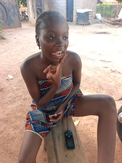 A young woman is sitting outdoors in Akwanga, Nigeria. She has dark skin and dark hair, is wearing an orange, blue, and white patterned garment. She is making a gesture with her right hand, and her tongue is slightly protruding. The woman sits on a wooden plank. A black feature phone is resting on the plank next to her thigh. The ground is reddish-brown dirt. In the background, there is a building made of cinder blocks, a metal gate, and vegetation. A motorbike is partially visible behind the gate.