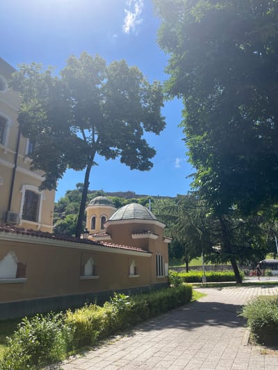 The image shows a sunny day in Plovdiv, Bulgaria. In the foreground, a paved walkway leads through a garden area with manicured bushes. To the left is a building with yellow walls and red-tiled roofs, featuring arched windows. Adjacent to this building is a smaller structure with a dome, also with arched windows. In the background, a hill rises, topped by a fortress. Trees with lush green foliage are scattered throughout the scene, with some casting shadows on the walkway. The sky is clear blue with a few wisps of white clouds. In the middle ground, a person is walking a dog on the paved area. The overall impression is one of a peaceful, historic urban environment bathed in sunlight.