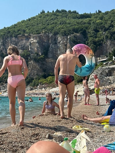 In Mirište, Montenegro, a group of people enjoy a sunny day at a pebble beach. The scene is dominated by a steep, green, tree-covered cliff face on the left, meeting a clear blue ocean on the right. Several individuals are visible, some standing on the beach, others partially in the water.
In the foreground, a person wearing a pink bikini with ruffled details stands facing away from the camera, their back and legs visible. To their left, the blue water of the sea laps at the shore. Further back, on the pebbly beach, a man in patterned swim trunks holds a colorful inflatable ring featuring palm trees. Nearby, another man in green swim trunks stands. A woman with dark hair wearing a patterned bikini is also visible on the beach.
In the background, more people are scattered along the beach, with some relaxing on loungers. The water is dotted with swimmers. The overall atmosphere is one of leisure and enjoyment, suggesting a warm, bright day. The sunlight casts shadows, indicating it is likely midday. No visible text is present in the image.
