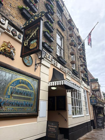 The image shows a street view of a brick building in London, United Kingdom, adorned with hanging flower baskets. A black sign with gold lettering reads "THE Copenhagen". Below this, a large, ornate framed sign with blue and gold lettering states "OUR FAMILY RESTAURANT". To the right, a smaller sign for "THE YACHT" restaurant is visible, hanging from the building. Several other signs and plaques are also present on the facade. A British flag flies from a flagpole on the building. The street appears to be paved, and the sky is overcast. There are no people visible in the image.