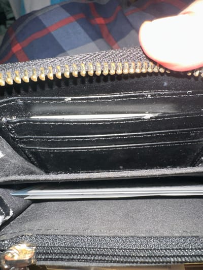 This close-up, slightly overhead image captures the opened interior of a black zippered wallet or purse, held by a person whose thumb is visible in the upper left corner. The wallet features multiple vertical card slots, made of a smooth, dark material, likely synthetic or leather. Several card slots are visible, mostly empty, though the white edge of at least one card is discernible within a slot towards the right.
Small white specks, resembling dust or minor debris, are scattered across the black surfaces of the card slots and along their seams, indicating typical wear and usage. A prominent metal zipper with gold-colored teeth and a dark fabric tape runs along the left edge of the wallet. Further inside, to the right, another internal zippered compartment is visible, with a metallic zipper pull partially showing. The internal lining of the wallet appears to be a dark, fine-textured fabric.
In the blurred background, a pattern of red and blue plaid, consistent with a shirt, can be seen. The lighting is bright, creating a significant highlight on the person's thumb. The activity depicted is the holding and opening of the wallet, suggesting an interaction with its contents or an examination of the item. There are no explicit location cues, time of day, weather, or discernible emotions. No visible text is present in the image.