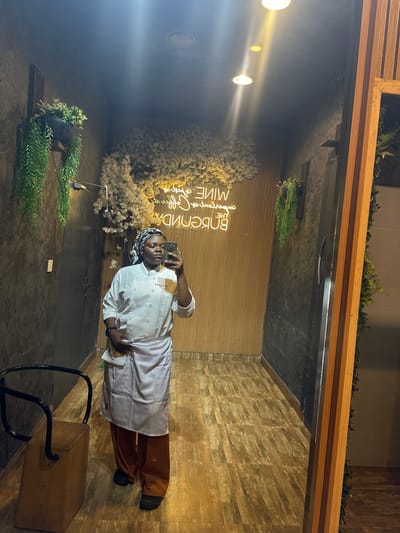 A full-body, medium shot captures a woman dressed in a white chef's uniform and orange pants, taking a mirror selfie in what appears to be a restaurant or bar. She is standing in a hallway with textured, dark walls and a wooden floor. Hanging plants adorn the walls, and a neon sign with text in a cursive font is visible in the background. A wooden chair with a black metal frame sits to her left. The lighting is dim, with spotlights illuminating the scene. The woman is looking at her phone, holding it in her right hand. The overall environment suggests a modern and stylish establishment.