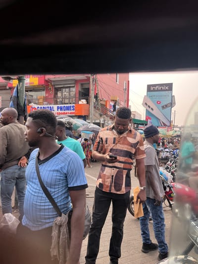 The image depicts a bustling street scene in Tudu, Accra Central, Ghana. The foreground is partially obscured by a dark horizontal bar, suggesting the photo was taken from a vehicle or enclosure.

Three men are prominent in the mid-ground. On the far left, a man wearing a light blue vertical-striped polo shirt with black trim and a black cross-body bag has a black bone-conduction earpiece on his left ear. He is facing left, partially out of frame. To his right and slightly forward, a man in a brown, black, and white abstract-patterned short-sleeved button-up shirt and dark trousers stands looking down at a black smartphone in his hands. He wears a white watch on his left wrist. To his right, another man wears a grey t-shirt, blue distressed jeans, a black beanie, and black shoes, holding a small yellow envelope or bag in his right hand. He stands near a red and black motorcycle.

The background reveals a dense urban environment with multiple structures. A red/pink building with "CAPITAL PHONES" prominently displayed on an orange sign is visible to the left, along with the phone number 0244641488. Overhead, numerous electrical wires crisscross. To the right, a large billboard for "Infinix HOT 60 SERIES" is visible. The street features numerous other pedestrians, market stalls with umbrellas, and additional motorcycles, indicating a busy commercial area. The sky appears overcast.
