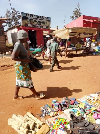 The scene takes place in Lwandeti, Kenya, at coordinates 0.563 latitude and 34.806 longitude. A woman in a grey shirt and a patterned skirt is walking down a dirt road. She is carrying a black plastic basin. In front of her are various items for sale including sandals, brooms, and kitchen utensils. There are also various stalls and shops, including a barber shop called "Fades Master". The barber shop is red and has a sign with a phone number. There is a motorcycle parked in front of the shop. There is another woman walking towards the right of the image, along with two other men. It appears to be a sunny day.