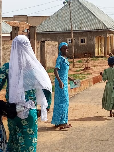 In Tempure, Nigeria, three young females are captured walking along an unpaved road lined with reddish-brown dirt. The scene is dominated by a single-story building with a gray, corrugated metal roof and plastered walls. Two windows with blue frames are visible on the building's exterior.
On the left, a female is partially visible, wearing a green dress adorned with a floral pattern and a white veil covering her head. She is facing away from the camera and appears to be walking.
To her right, a female dressed in a vibrant blue, patterned long-sleeved dress and a matching headscarf is walking towards the camera. She is holding a small, colorful booklet or magazine in her right hand. Her gaze is directed at the camera.
Further to the right, a third female, seen from the back, is wearing a green dress and a dark head covering. She is also walking away from the camera.
The environment consists of dry, dusty ground with sparse green vegetation. A thin, utility pole stands to the left of the building. The sky is overcast.