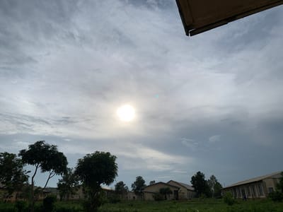 The image captures an outdoor scene in Kuru Karama, Nigeria. The sky is overcast with scattered clouds and the sun is visible as a bright, diffused orb in the upper center of the frame, casting a soft light. In the foreground, there are several trees with dense foliage, some appearing to be mango trees. Behind the trees, a series of light-colored, single-story buildings with sloped roofs are visible. These structures have numerous windows. The ground is covered in lush green grass and some smaller plants. A section of a wooden structure is visible in the upper right corner, appearing to be a roof overhang. The overall impression is one of a rural or semi-rural setting, possibly a school or an institutional compound.
