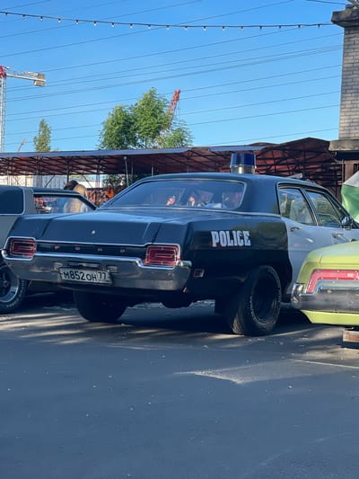 The image captures a vintage black and white police car parked outdoors. The car features a blue light bar on its roof and "POLICE" written in white on its side. The license plate reads "M852OH 77 RUS". In the background, there is a building with brickwork and a canopy structure. A bright blue sky is visible above, with some power lines and a red crane in the distance. The ground appears to be asphalt. The overall scene suggests an outdoor event or exhibition, possibly in Saint Petersburg, Russia, given the license plate format.