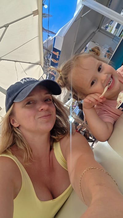 A blonde woman wearing a yellow tank top and a navy blue baseball cap is in the foreground of the image, looking at the camera with a slight smile. To her right, a young blonde girl with pigtails is eating ice cream and looking at the camera with a wide smile. They are seated at a table outdoors, likely at a cafe or restaurant. In the background, there are umbrellas and structures that suggest a tourist area. The lighting indicates it is daytime, with bright sunlight. The overall scene appears to be relaxed and casual. The location is identified as Puerto De Santiago, Spain.