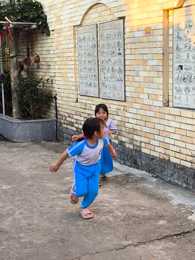 The scene, located in Thành phố Quảng Ngãi, Vietnam, depicts two young girls outdoors on an uneven, cracked concrete surface adjacent to a building. The foreground features a girl wearing a white short-sleeve shirt with blue trim on the collar and sleeves, blue pants, and pink open-toe sandals. She is in motion, captured mid-stride with her left leg forward, arms slightly extended away from her body, and head turned towards the viewer's right. A pink band is visible on her left wrist. Behind her, a second girl, similarly dressed in a white short-sleeve shirt with blue trim, a blue skirt, and brown sandals, stands facing generally towards the foreground girl, exhibiting a smile.
The background consists of a building with a light tan brick wall featuring a darker grey base. Three large, vertically oriented, framed posters are mounted on the wall. The two left posters are titled "TRANH THUYET BIEU" with the English subtitle "Illustrations for the Law of Karma," while the right poster is titled "TRANH NHAN QUA" with the English subtitle "Illustrations for the Law of Cause and Effect." Each poster contains grids of small black and white illustrations. An arched brick design is present above the two left posters. Between the middle and right posters, a small orange sticker with text and the phone number 0905466333 is affixed to the wall. To the far left of the frame, a grey concrete planter box contains green leafy plants and a small shrub with red blossoms, with additional dense foliage overhanging from above. A dark vertical utility pole or pipe structure is visible near the planter. The upper right portion of the brick wall is illuminated by a distinct, warm yellow light, indicative of direct sunlight.