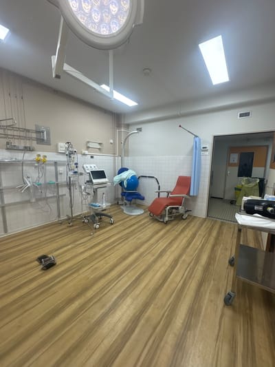 The image depicts the interior of an operating room, likely located in a hospital or medical facility in Tourcoing, France. The room features medical equipment, including a surgical light fixture, patient monitor, and medical gas outlets. A medical chair and hospital beds are also present. The walls are tiled, and the floor is covered with wood-patterned linoleum. A doorway leads to another room, visible in the background. The environment appears sterile and clinical. There are no people visible in the image.