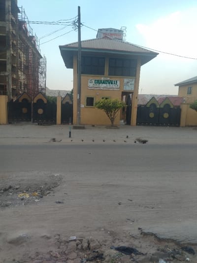 The image shows a street in New Karu, Nigeria. The street is paved with asphalt, but there are several potholes and cracks in the surface. There are several pieces of trash and debris scattered along the edges of the road. In the foreground, there is a small tree. The scene is captured from a street level perspective. There is a building in the background. The building is a tan color and appears to be a hotel or guest house. The building has a sign that reads "Grandville Rest House," "Reasonable Accommodation." The sign is located above a gate with a sign that reads "Entry." There is also a water tank on the roof. The building has several windows and a large, open-air space on the second floor. The building has several windows and a large, open-air space on the second floor. There is a chain-link fence in front of the building. There is a small tree in front of the building. The scene is taken on a cloudy day with a bright blue sky visible. The image was taken from a street level perspective. There are no people in the image. The only subject is the Grandville Rest House building. The image suggests that the building is located on a street with moderate traffic.
