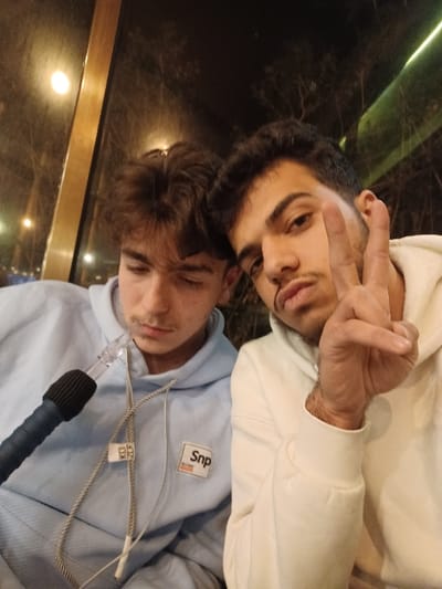 Two young men pose for a selfie. The top man, wearing a white hoodie, is making a peace sign with his fingers. The other man, below, is wearing a light blue hoodie with a "Snp" patch on the chest and appears to be holding a hookah. The background is a dimly lit, blurry scene, possibly outdoors with greenery and a window. The image is slightly rotated.