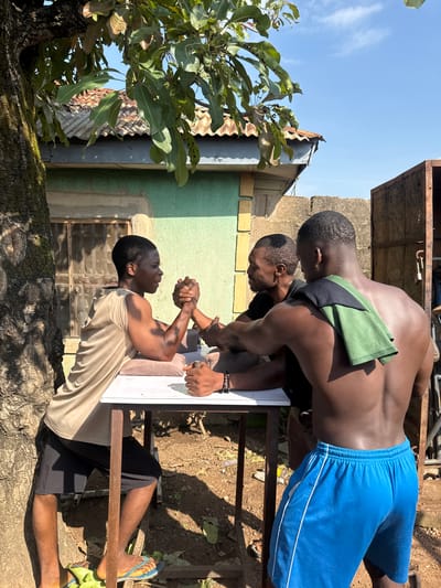 Three Black men are engaged in an arm-wrestling match outdoors. Two men are positioned opposite each other at a table, their arms clasped, in the midst of their competition. The third man stands behind one of the competitors, observing the match. The scene is set in Akwanga, Nigeria, indicated by the architectural style of the buildings and the surrounding vegetation. The background features a green-painted building with a corrugated metal roof and a large tree on the left, casting shadows on the ground. The ground is dirt, scattered with fallen leaves. The sky is clear and blue.