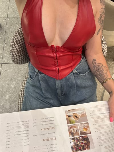 The image is a close-up, waist-up shot of a person seated at a table in what appears to be a cafe or restaurant. The person is wearing a red leather halter top and denim shorts. They have tattoos on their arm, including a snake design. A menu is open on the table in front of them, displaying a list of drinks and desserts. The background shows a tiled floor and part of a chair. The overall impression is that of a casual dining or social setting, likely indoors given the lighting and lack of outdoor elements. The menu has text in a language that appears to be Serbian, based on the word "Kombucha" and the Cyrillic script for "Pivo/Beer". This suggests the location is in Belgrade, Serbia.