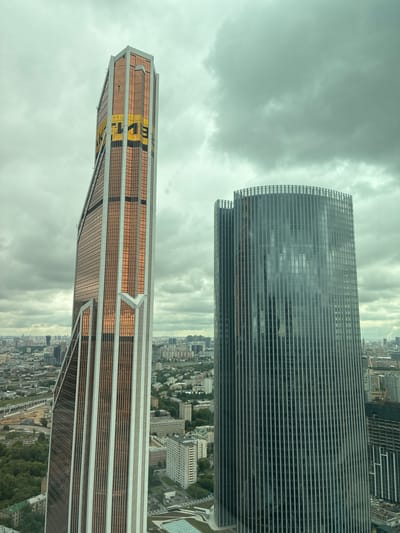 An aerial, eye-level, wide shot captures two skyscrapers against a cloudy, overcast sky. The skyscraper on the left is tall and angular, with a bronze-tinted glass facade and white vertical accents. Large yellow lettering, partially obscured by the angle, is visible on its upper section. The skyscraper on the right is cylindrical with a dark, reflective glass facade, featuring thin vertical silver accents. Both buildings dominate the frame, with a sprawling cityscape visible in the background, including smaller buildings, trees, and infrastructure. The lighting is diffused due to the clouds, creating a muted color palette. The image appears to be taken from a high vantage point, possibly another building or a drone. The context provided indicates the location is in the city of Khimki, Russia.
