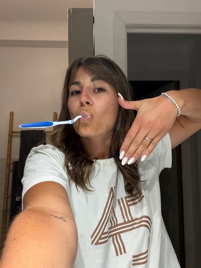 A woman is taking a selfie while brushing her teeth indoors. She is wearing a white t-shirt, a bracelet, and a ring. The background shows a window and a bamboo chair. There is no visible indication that the location is Cannes, France.