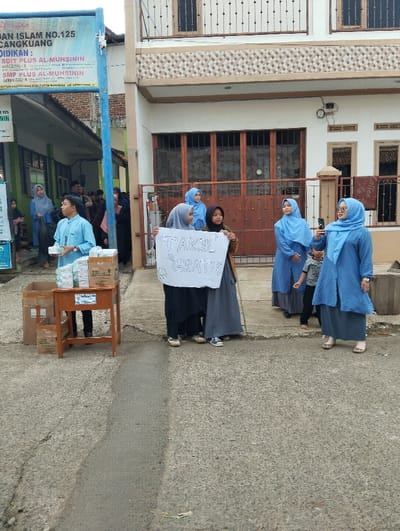 The image shows a group of five young women in blue and grey clothing standing in front of a two-story house in Cangkuang, Indonesia, located at latitude -7.065, longitude 107.552. The women are holding a white sign with the words "TAKLIF SYARAT" written on it. There are a few boxes of unknown contents to the left, and a table with a few boxes of unknown contents on it. The house is made of concrete and has a metal fence. There are several windows and doors. The scene is daytime.
