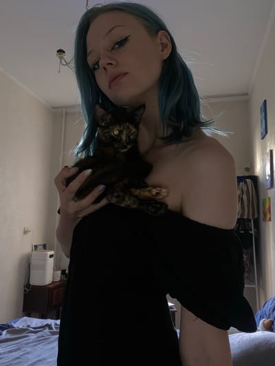 The media file shows a close-up, vertically oriented, selfie-style image of a person holding a tortoise-shell cat. The person has blue hair and is wearing a black off-the-shoulder top. Their face is partially visible, with makeup including eyeliner and what appears to be drawn-on beauty marks. The cat is nestled against the person's chest, and its paws are visible. The background suggests an indoor setting, possibly a bedroom, with a wall, a ceiling with some artwork, and furniture. A white appliance, possibly a heater or humidifier, is visible on a shelf. The lighting suggests it is likely daytime, but the room is not brightly lit. The overall mood is intimate and relaxed, with the person and cat appearing comfortable together.