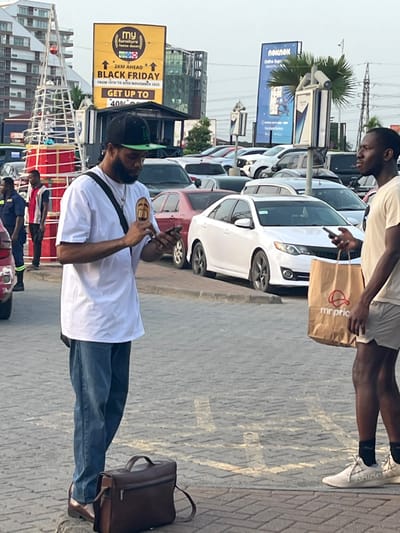 This image depicts an outdoor urban scene in Accra, Ghana, likely a busy street or commercial area. In the foreground, two men are visible on a paved surface. On the left, a man with a beard, wearing a white shirt, blue jeans, and a green baseball cap, is focused on his mobile phone held in both hands. A dark brown shoulder bag hangs across him, and a matching briefcase rests on the ground nearby. To his right, another man, dressed in a light-colored t-shirt, shorts, and white Adidas sneakers, holds a smartphone in one hand and a brown paper shopping bag with the "Mr Price" logo in the other.

Behind the individuals, a line of parked cars, predominantly white sedans, stretches along the street. The background features various urban elements including buildings, streetlights, and power lines. Two prominent billboards are visible: one yellow billboard on the left advertises "my furniture" with text promoting "BLACK FRIDAY FROM 15TH TO 25TH NOVEMBER 2025 GET UP TO 40%". Another blue billboard further back features "Nokia Online Store". Several other people, some in uniforms, can be seen in the distance, indicating a populated area. The scene is brightly illuminated by daylight under a clear sky, suggesting a sunny day. The overall impression is that of a typical, active urban environment.