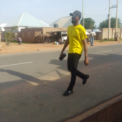 The image depicts a street scene in Tempure, Nigeria. In the foreground, a man wearing a yellow t-shirt, black cap, glasses, black pants, and black sneakers is walking towards the right side of the frame. He is holding a dark-colored object, possibly a phone or small book, in his right hand. A watch with a red face is visible on his left wrist. His shadow is cast on the asphalt road.
In the background, across the street, several buildings are visible. One building on the left has a distinctive pyramid-shaped light-colored roof. Next to it is a building constructed with mud bricks. Further in the background, there are more buildings and trees. Two individuals are on a motorcycle in the left background. Another person, a man wearing a light-colored t-shirt and blue jeans, is standing near a stall in the mid-ground. A woman is seated behind a stall. Power lines and utility poles are visible on the right side of the image. The road is paved with asphalt and has a white line marking. The overall environment appears to be a rural or semi-urban area with a clear sky.