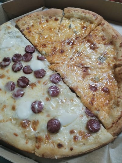 In Mykolaiv, Ukraine, a pizza sits in a cardboard box. The pizza is divided into four slices, with two distinct toppings. One half of the pizza is topped with sliced sausages and white cheese. The other half is topped with melted golden cheese, some of which is browned and slightly crispy. The pizza is in a brown cardboard pizza box.