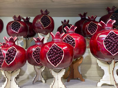 A close-up, eye-level shot captures a collection of decorative pomegranate figurines displayed on wooden stands. The pomegranates are a deep, glossy red, with intricate carvings revealing an inner core of dark red seeds. The figurines vary in size and are arranged in multiple rows on a light-colored, horizontally paneled surface, likely a shelf. The background consists of similar shelving, suggesting a retail or display setting. The lighting is bright and even, highlighting the texture and sheen of the decorative objects. There are no people visible in the image. This scene is from a souvenir shop in Sochi, Russia, where such crafts are popular gifts for tourists.