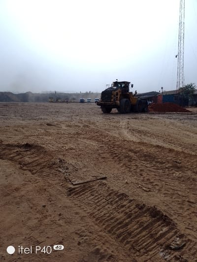 The image depicts a large, open construction or industrial site located in Hoche, Nigeria, under a uniform, overcast sky. The ground consists of reddish-brown soil, extensively marked by fresh, heavy tire tracks indicating recent vehicle activity, with several areas appearing muddy or damp. In the midground, a prominent yellow wheel loader is parked, positioned towards the right side of the frame, adjacent to a distinct pile of reddish-brown earth. Behind this pile, a blue shipping container or temporary structure is visible. Further to the right and extending high into the background, a tall, metallic lattice communication tower is present, with some sparse green foliage at its base. In the distant background, a line of stationary vehicles, including at least three yellow dump trucks and several lighter-colored trucks, is discernible. To the far left background, a large, dark mound of material, consistent with aggregate or excavated earth, is present, partially obscured by atmospheric haze. A single, indistinct human figure is visible walking or standing in the far background, near the line of vehicles. The scene conveys an active or recently utilized site for heavy machinery operations.