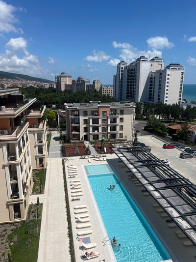 This aerial perspective captures a vibrant scene on a sunny day in Nesebar, Bulgaria. The primary focus is a rectangular swimming pool filled with clear blue water, where several people are enjoying a leisurely swim. Loungers are neatly arranged on the surrounding tiled area, with some unoccupied and others occupied by individuals relaxing.

To the left of the pool, a multi-story building with a black metal fence runs along its edge. Beyond this, a parking lot is visible, with several cars parked, including a striking red one. Further in the distance, a cluster of modern, light-colored apartment buildings with numerous windows and balconies dominate the skyline, interspersed with lush green trees. The architecture suggests a resort or residential complex.

The background reveals a vast expanse of the sea under a bright blue sky dotted with fluffy white clouds, hinting at a warm, pleasant day. The overall impression is one of a relaxed holiday atmosphere. The image is oriented vertically, requiring a rotation to view the scene as intended. No discernible text is present in the image.