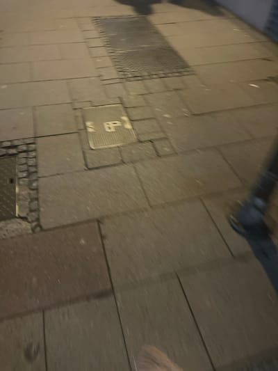 A textured gray pavement, composed of interlocking stones, is visible. A metal grating, square with small holes, sits within the pavement. Another square metal plate, marked with an upright "P," is visible. A section of gray piping runs along the pavement's upper edge. At the bottom, another metal square with textured lines is placed inside the interlocking stones. A partial blurry beige object appears at the upper right.