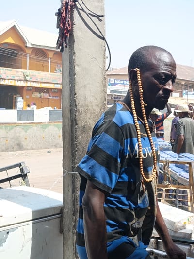 The image depicts an outdoor urban scene in Fagge C, Nigeria, under bright, clear daylight. In the foreground, a dark-skinned adult male with a shaved head is positioned on the right side of the frame, facing slightly left and downward. He wears a blue and black horizontally striped short-sleeved t-shirt, exhibiting minor wear and small holes on the sleeves. A long string of light brown, spherical wooden beads is looped over his right ear, resting against his neck and chest.
Adjacent to the man, centrally positioned, is a rough, grey concrete utility pole with several dark electrical wires and strips of red fabric attached near its top. A thin white wire extends horizontally from the pole towards the lower left. Partially visible in the lower left foreground is a weathered, white chest freezer with its lid closed.
In the background on the left, a two-story building with a light orange facade and white roof features arched openings on the upper level and commercial signage, including text such as "KIVQ" and "LOKACI MAI KYAU". A low white and dark grey barrier runs horizontally below this building. The ground beyond appears dusty. In the right background, multiple individuals are present, one wearing a striped shirt and a light hat, and another in a dark top and a straw hat. A wooden cart laden with stacks of rectangular light blue packages is visible among these figures. Further back, a building with a sign reading "MAZAF SHO" is partially visible. The sky is a clear, light blue.