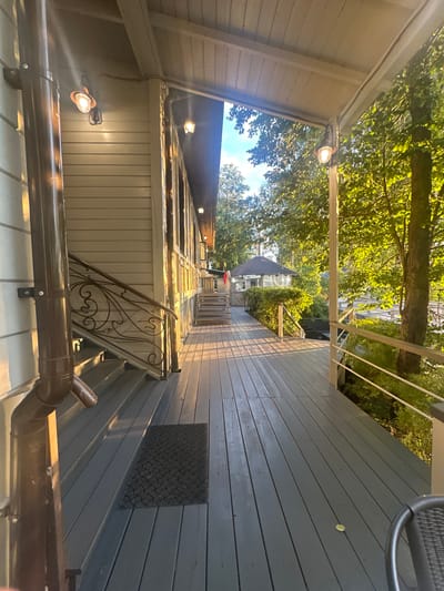 The image depicts a covered wooden deck and staircase of a building, likely a house or a resort, situated in a natural setting with trees surrounding it. The perspective is from the deck, looking towards the stairs and then further into the distance where a gazebo or small pavilion is visible amongst the foliage. Sunlight filters through the leaves, casting dappled shadows on the deck and stairs. There are outdoor lights attached to the building and on posts along the deck. A brown downspout runs along the side of the building next to the stairs. A dark doormat is placed on the stairs. There are no people clearly visible in the image. The environment suggests a peaceful, outdoor recreational space. The image does not provide any explicit clues to its location within Moscow, Russia.