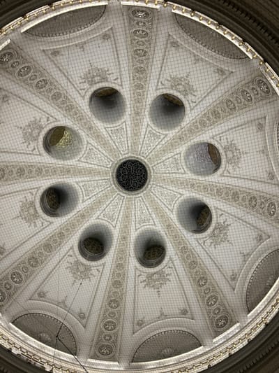 The image shows an interior view looking directly up at the ornate dome of the Austrian National Library (Österreichische Nationalbibliothek) in Vienna, Austria. The dome features intricate white plasterwork with radiating patterns, decorative friezes, and a central circular opening with a decorative metal grille. Surrounding the central opening are eight smaller circular apertures, through which light and gilded decorative elements are visible. The entire dome is covered by a fine mesh netting, likely for safety or protection. The surrounding architecture includes curved sections and a golden decorative railing at the top edge of the visible dome structure. There are no people or visible actions occurring in the scene.