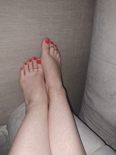 The image shows a close-up view of a person's feet and lower legs resting on a neutral-toned textured surface, likely a sofa or upholstered furniture. The feet are crossed, with the toes pointing upwards and slightly towards the viewer. The toenails are painted a bright red color. A simple silver ring is visible on the second toe of the left foot. The skin on the legs and feet is fair and has numerous freckles. The background consists of a textured, taupe-colored wall or fabric. The lighting is dim, casting soft shadows. There is no discernible indication of the location within the city of Chișinău, Moldova.